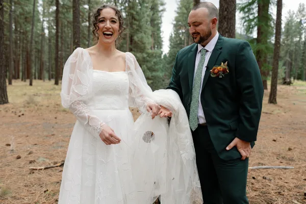Couple portrait of bride laughing in puff sleeve wedding dress and veil, holding hands with groom in green suit amid tall pines