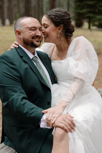 Couple portrait of bride and groom laughing as she hugs him in a forest clearing, her sheer puff sleeves and his green suit visible