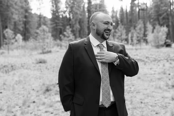 Groom portrait in a suit laughing candidly, showing his wedding ring as he stands among forest trees and meadow grass outdoors