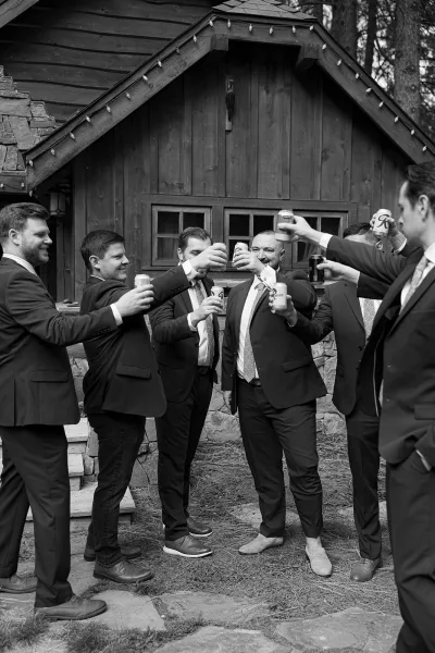 Groomsmen toast with beer cans, wearing matching suits and neckties on stone steps outside a wood cabin with string lights and trees