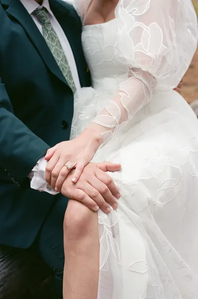 Couple portrait of bride and groom close up holding hands, highlighting wedding ring and her puff sleeve dress against blurred greenery