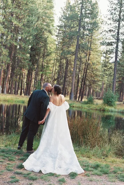 Wedding kiss as bride and groom hold hands by a pond in a pine forest, her off-the-shoulder dress train flowing behind