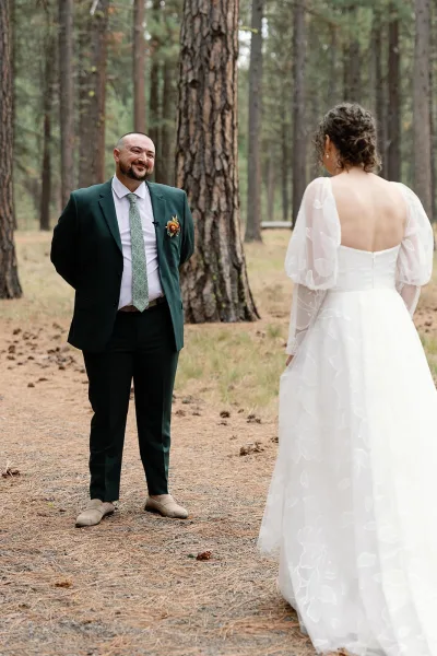 Wedding first look as bride in an open-back puff-sleeve dress approaches groom in a dark green suit on a pine forest path