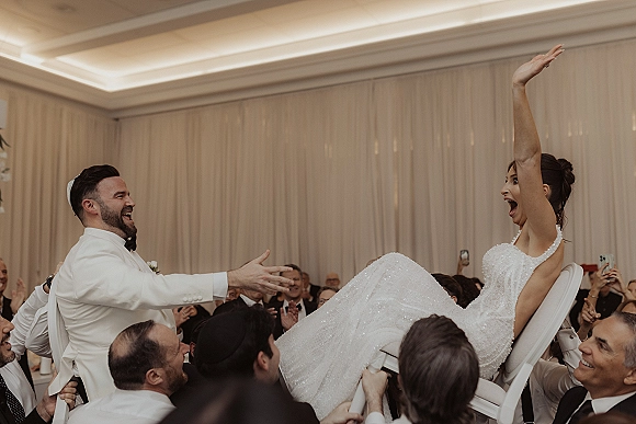 Hora chair lift at a Jewish wedding hora as bride in sparkly dress and groom in white tuxedo are raised on chairs amid cheering guests indoors