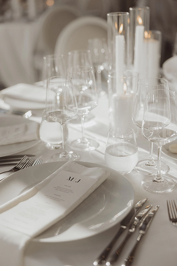 Reception tablescape with wedding place setting of white plates, folded napkins with menu card, glassware, silver flatware, and taper candles in glass cylinders