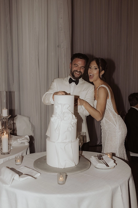 Wedding cake cutting as bride in beaded dress and groom in white tuxedo slice a two-tier cake at a candlelit reception table
