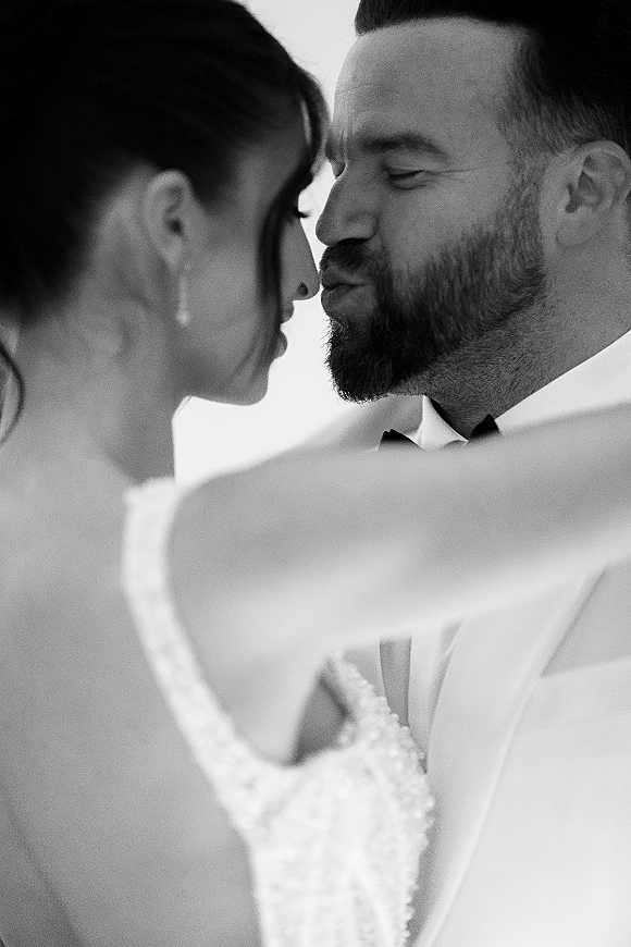 Wedding kiss portrait in black and white as bride in strapless lace bodice and pearl drop earrings kisses groom in bow tie by bright window light