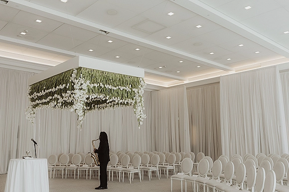 Ceremony setup with an indoor wedding ceremony chuppah with greenery, white florals, hanging canopy, and rows of white chairs in a draped ballroom