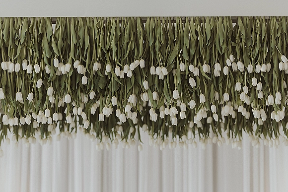 Hanging floral installation with white tulips and greenery garland cascading from a ceiling beam against soft white drapery backdrop