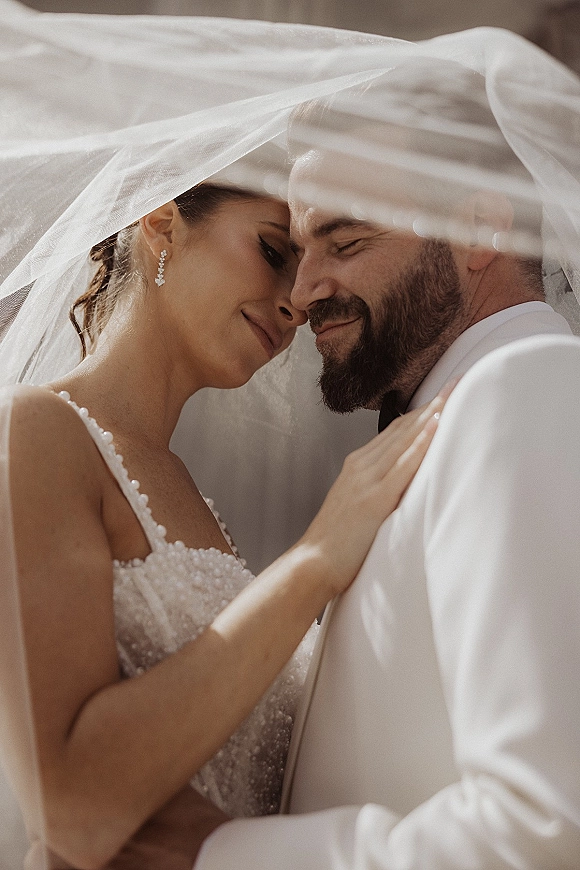 Couple portrait of bride and groom close up with a bridal veil draped over them, touching foreheads in soft natural light by a neutral wall