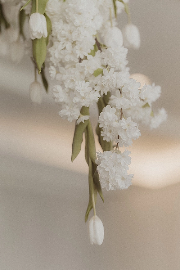 Hanging wedding flowers in a hanging floral installation of white tulips and greenery, softly lit by candlelight against a neutral wall