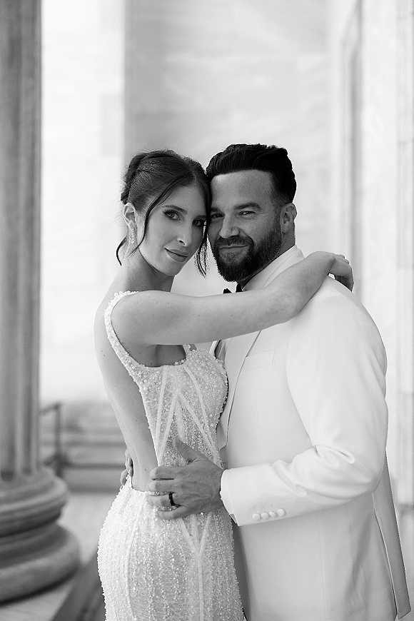 Couple portrait in a black and white wedding portrait style, bride in beaded gown with pearl straps embracing groom in bow tie by stone columns