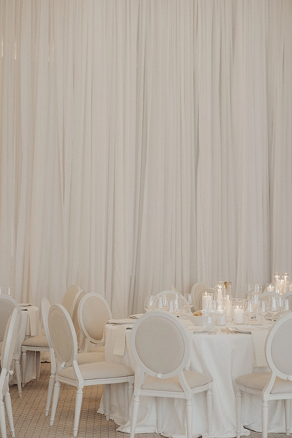 Reception tablescape with a white wedding tablescape of round tables, white linens, glass votives and pillar candles, set before draped curtains