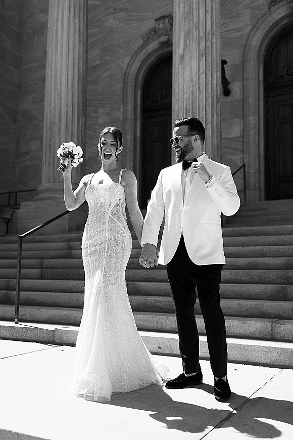 Newlywed couple portrait holding hands, bride with bouquet beside groom in white tuxedo and sunglasses on sunlit stone steps by columns