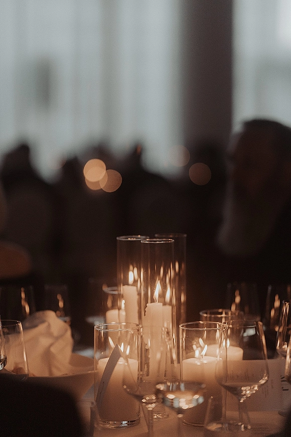 Candlelit tablescape with wedding candle centerpiece of white pillar candles in glass cylinders, wine glasses and place cards in a moody reception room