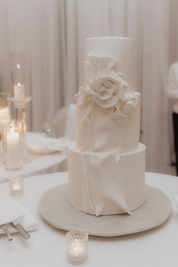 Wedding cake with fondant draping, a sugar flower accent, and votive candles on a linen banquet table against white drapery
