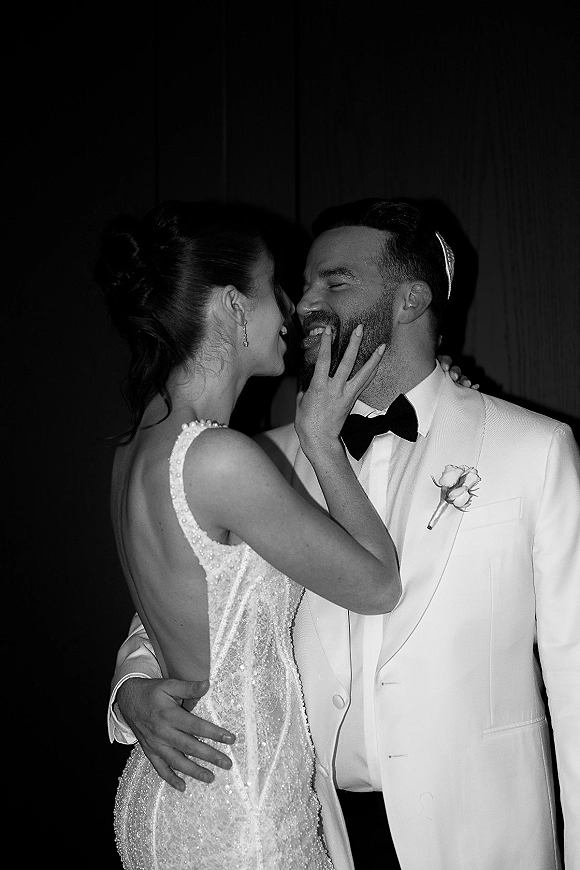 Wedding couple portrait, black and white wedding portrait of bride touching groom’s face as they laugh, against a dark curtain backdrop