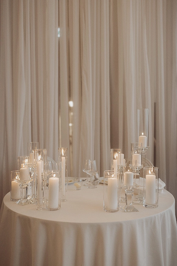 Wedding table decor with a candlelit wedding table featuring white taper candles in glass vases and wine glasses against neutral drapery curtains
