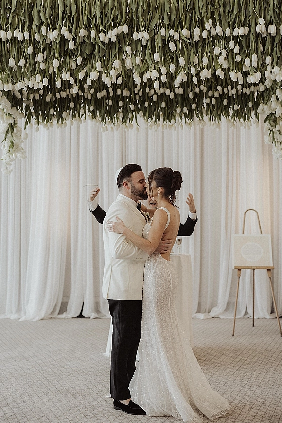 Wedding kiss as bride in a sparkly open-back gown embraces groom in a white jacket before white drapery and hanging flowers indoors