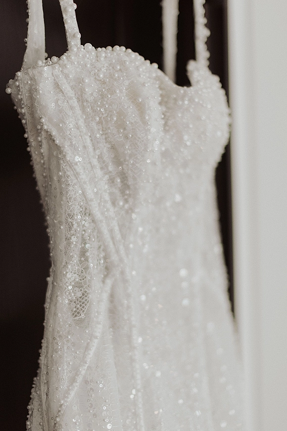 Wedding dress detail with pearl beaded bodice, sequins and lace fabric on spaghetti straps, hanging against a neutral wall with shadow