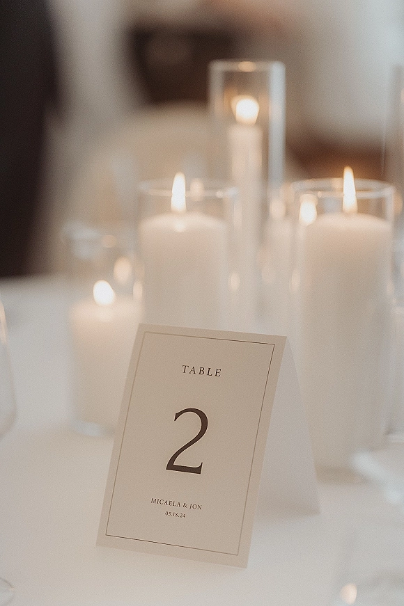 Wedding table number card with printed typography beside glass cylinder vases and pillar candles on a white tablecloth indoors