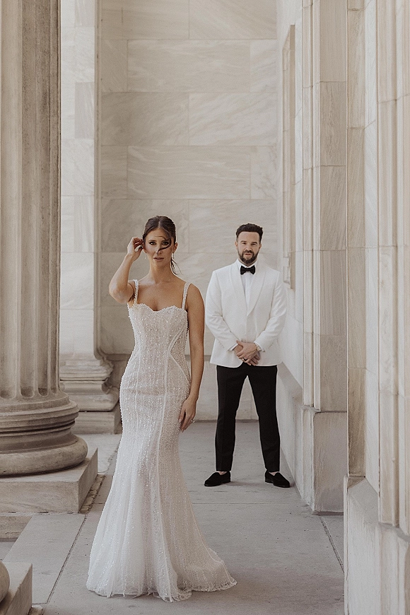 Couple portrait of bride in a beaded spaghetti-strap gown and groom in a white tuxedo jacket with black bow tie by stone columns