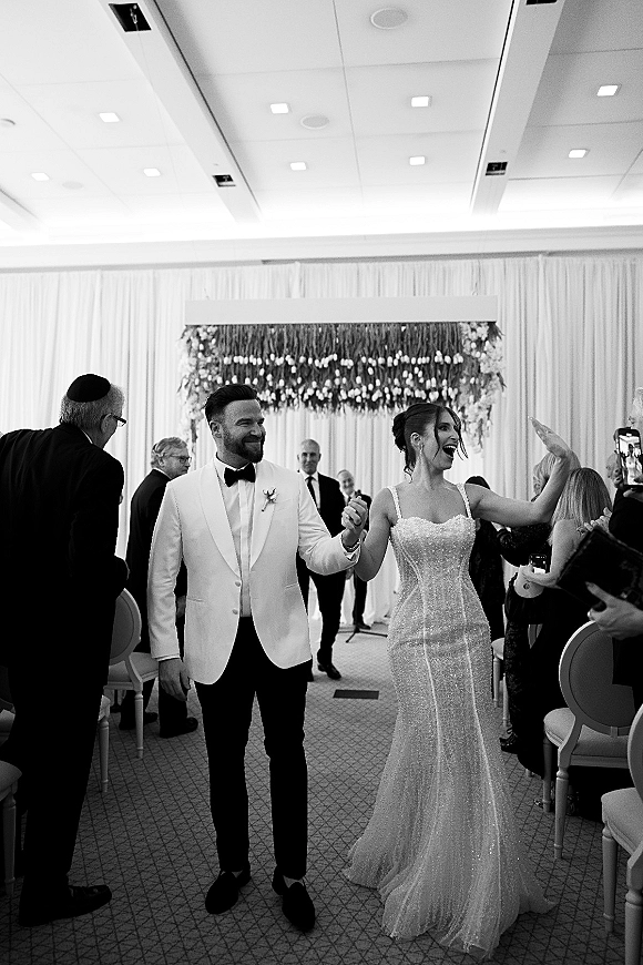 Wedding recessional as bride waves while holding groom’s hand, her sparkly strapless gown beside his white jacket under a floral chuppah indoors
