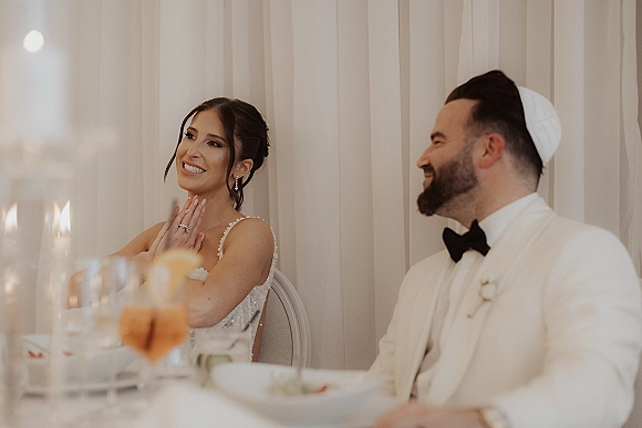 Reception speech as the bride giving toast in a beaded-strap gown beside groom in tux and kippah at candlelit head table with sheer drapery backdrop