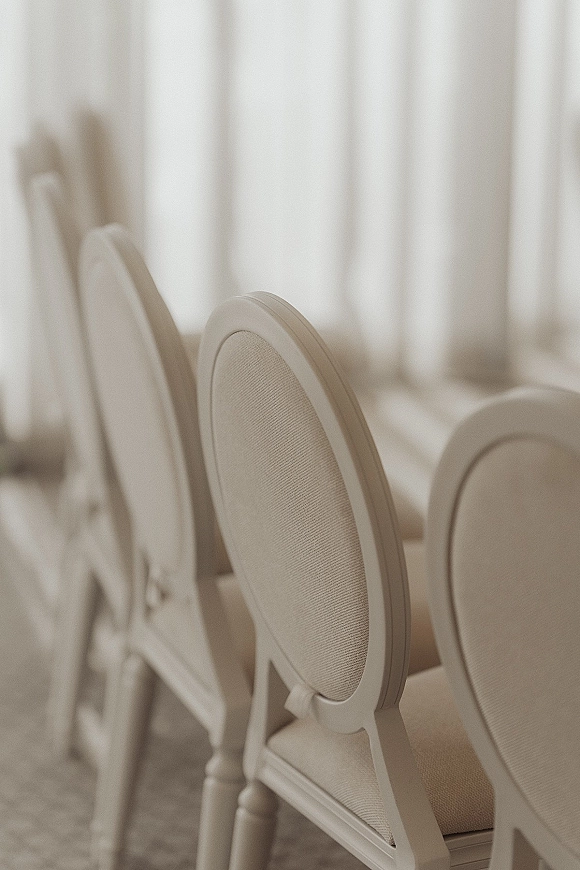 Wedding reception chairs in white upholstered oval-back style lined on a carpeted floor with soft white drapery behind