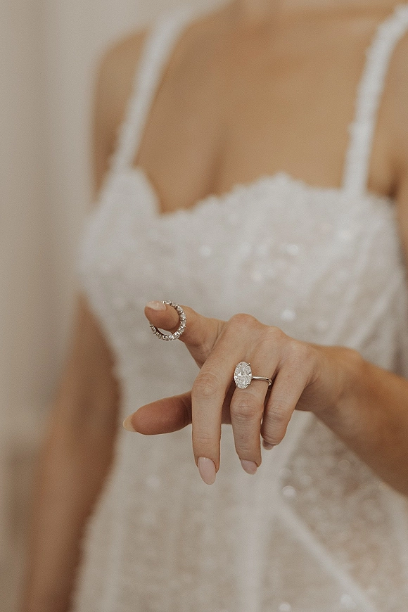 Engagement ring with an oval engagement ring diamond on manicured finger as the bride holds a hoop earring against a neutral backdrop