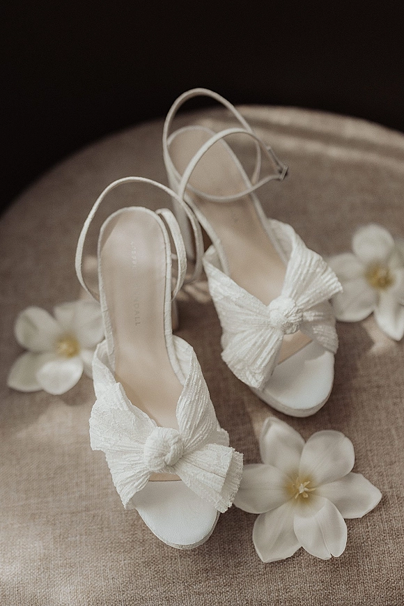 Bridal shoes with white bow heels and ankle straps styled beside white flowers on an upholstered chair with neutral fabric background
