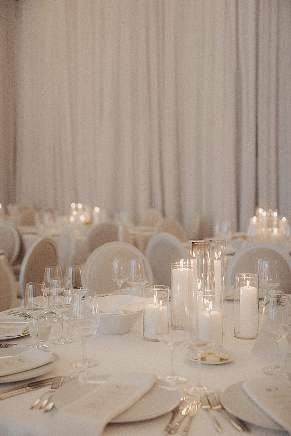 Reception tablescape with white wedding tablescape details, pillar candles in glass hurricane vases, crisp linens, and formal place settings under draped curtains