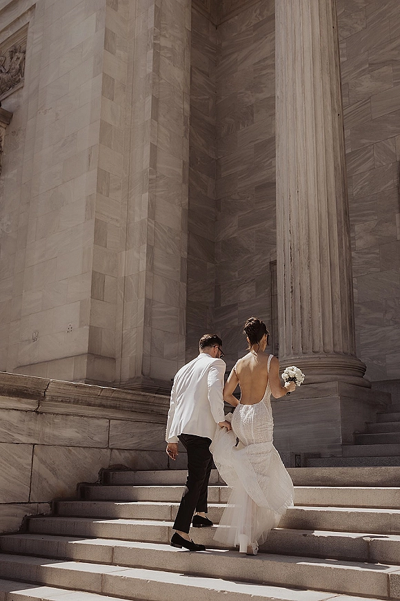 Couple portrait of bride and groom walking away up stone steps, bride lifting her train with bouquet, marble columns behind