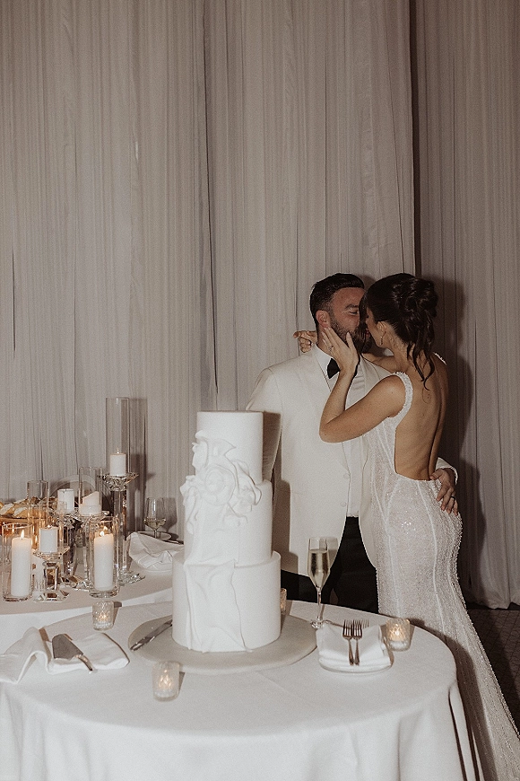 Wedding kiss portrait of bride and groom kissing beside a three-tier white cake on a candlelit table against draped curtains backdrop
