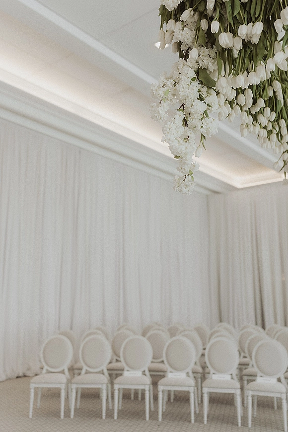 Ceremony setup for an indoor wedding ceremony with white tulip hanging florals above white chairs, framed by white draped walls