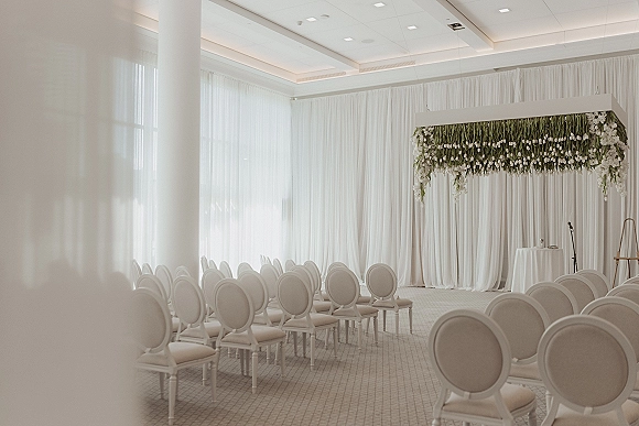 Indoor ceremony setup in a hotel ballroom ceremony with white drapery and floral chuppah, hanging greenery, and rows of chairs by tall windows.