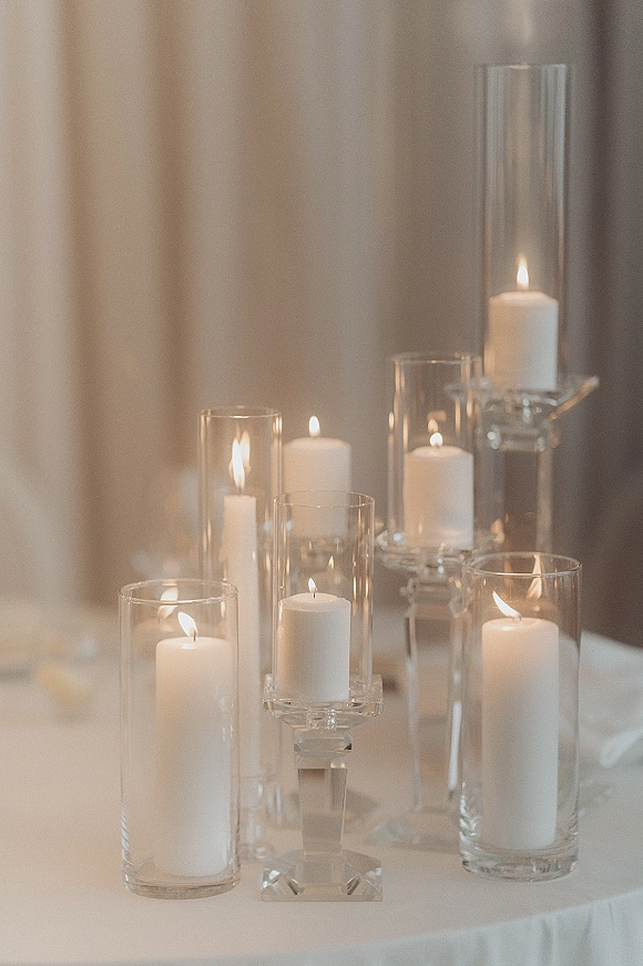 Wedding candle decor with white pillar candles in glass cylinder vases and clear pedestal stands against a draped fabric backdrop