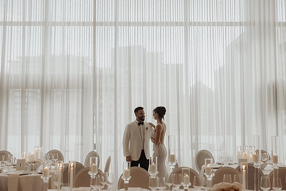 Couple portrait of bride and groom holding hands by floor-to-ceiling windows, city skyline behind, candlelit reception tables nearby