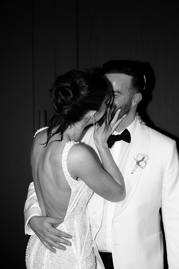 Wedding kiss as bride and groom embrace, her hand on his face, in front of a dark curtain backdrop at an indoor reception