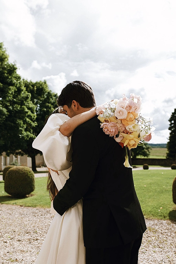 Wedding kiss as the bride and groom kissing, bride holding a rose bouquet with ribbon on a garden lawn beside trimmed hedges under cloudy sky
