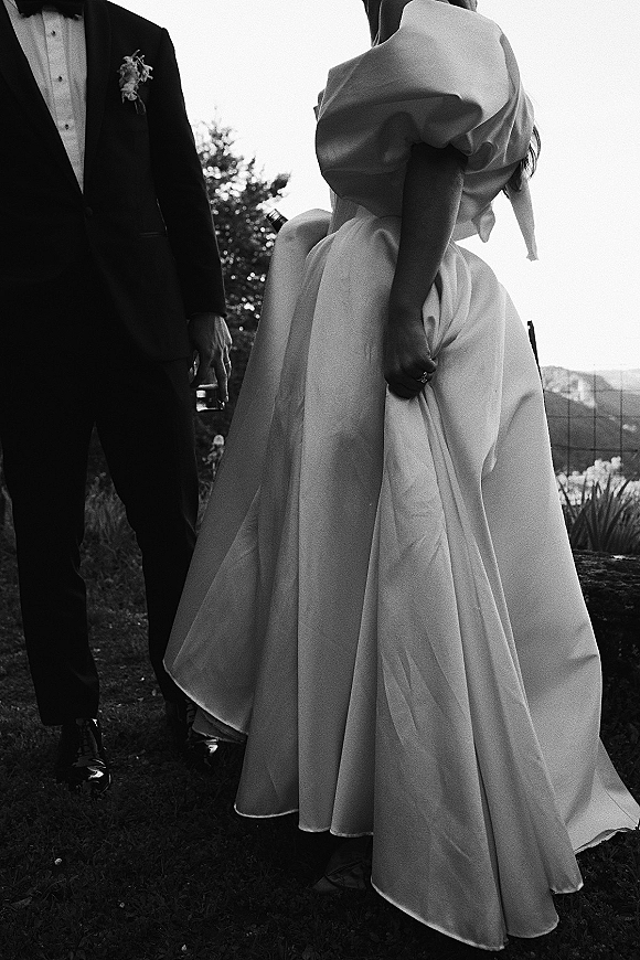 Couple portrait in a black and white wedding photo, bride in puff sleeve gown holding hem beside groom with wine bottle on a lawn hillside