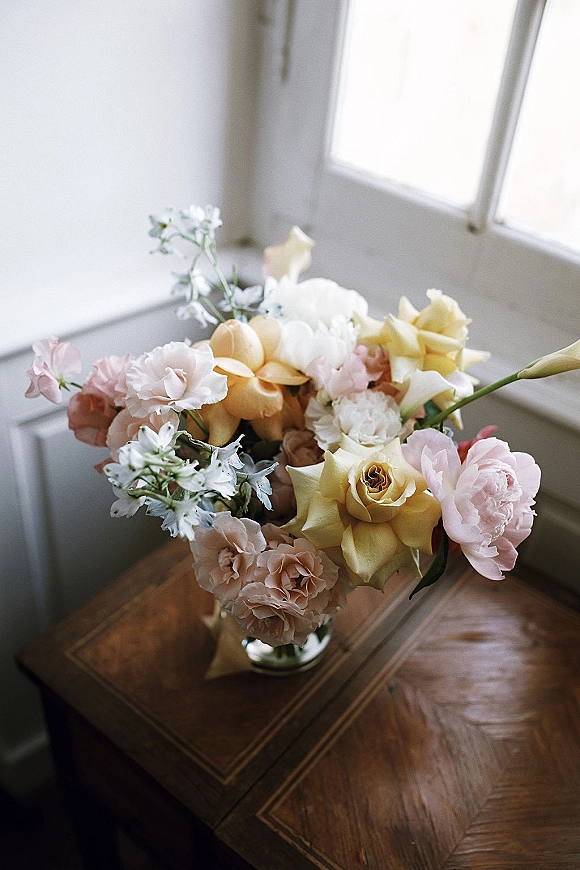 Wedding bouquet of roses, peonies, and carnations in a vase on a wooden table, softly lit by window light against a white wall
