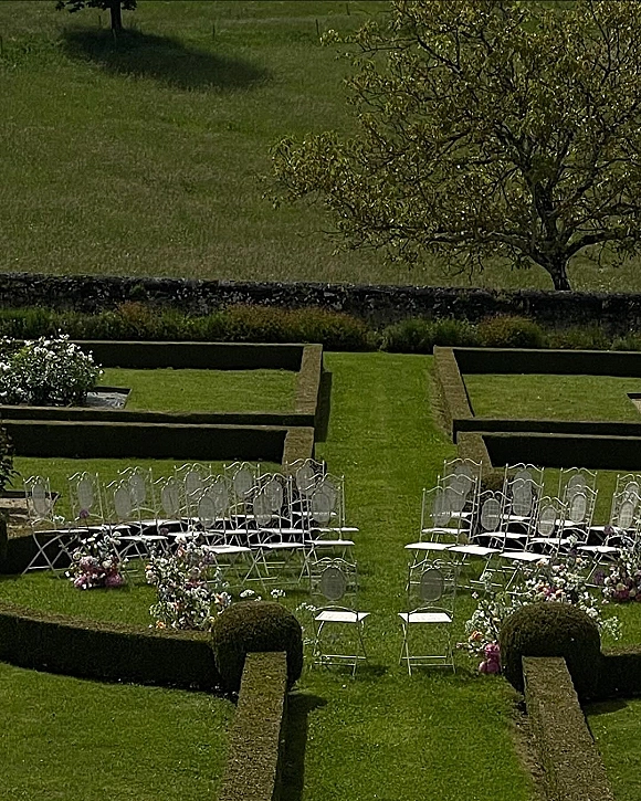 Ceremony setup with outdoor wedding ceremony seating, white metal chairs flanking a hedge-lined grass aisle with pink and purple florals in a terraced garden