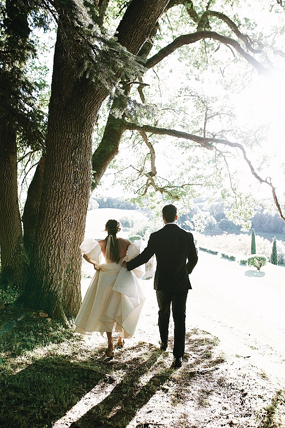 Couple portrait of bride and groom walking away hand in hand under a large tree, bride’s veil trailing in dappled sunlight