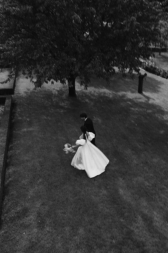 Couple portrait in a black and white wedding photo from above, bride twirling her dress with bouquet beside groom in suit on grass lawn near trees