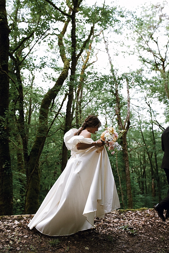 Bridal portrait of a bride holding a bouquet in an off-the-shoulder ball gown, looking down amid mossy forest greenery and trees