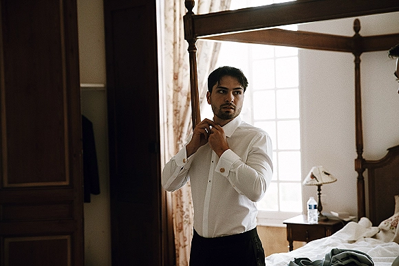 Groom getting ready as he buttons his white dress shirt, adjusting cufflinks beside the bed in a naturally lit bedroom by the window