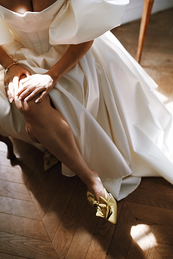 Bridal shoes in gold wedding heels with satin bow, as the bride adjusts them on a wood floor beside a chair, ringed hand visible