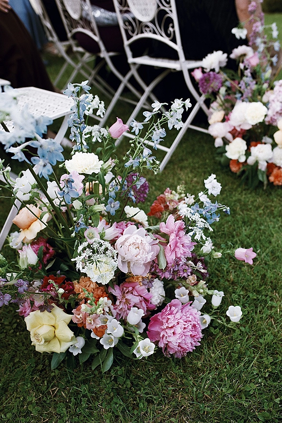 Aisle florals with ground aisle flowers in pastel clusters of garden roses, peonies, and blue delphinium beside white metal chairs on a lawn