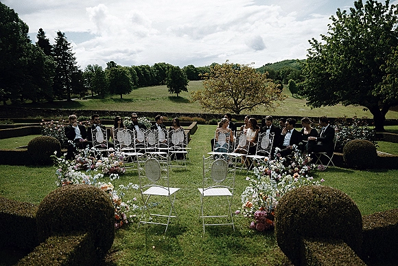 Ceremony seating with white wrought iron chairs and flower-lined wedding aisle, guests in tuxedos on a manicured lawn under cloudy sky